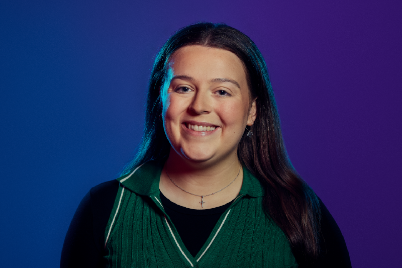 Headshot of a dark haired young female wearing a green v-neck  over a black long slieved shit, standing in front of a blue to purple background